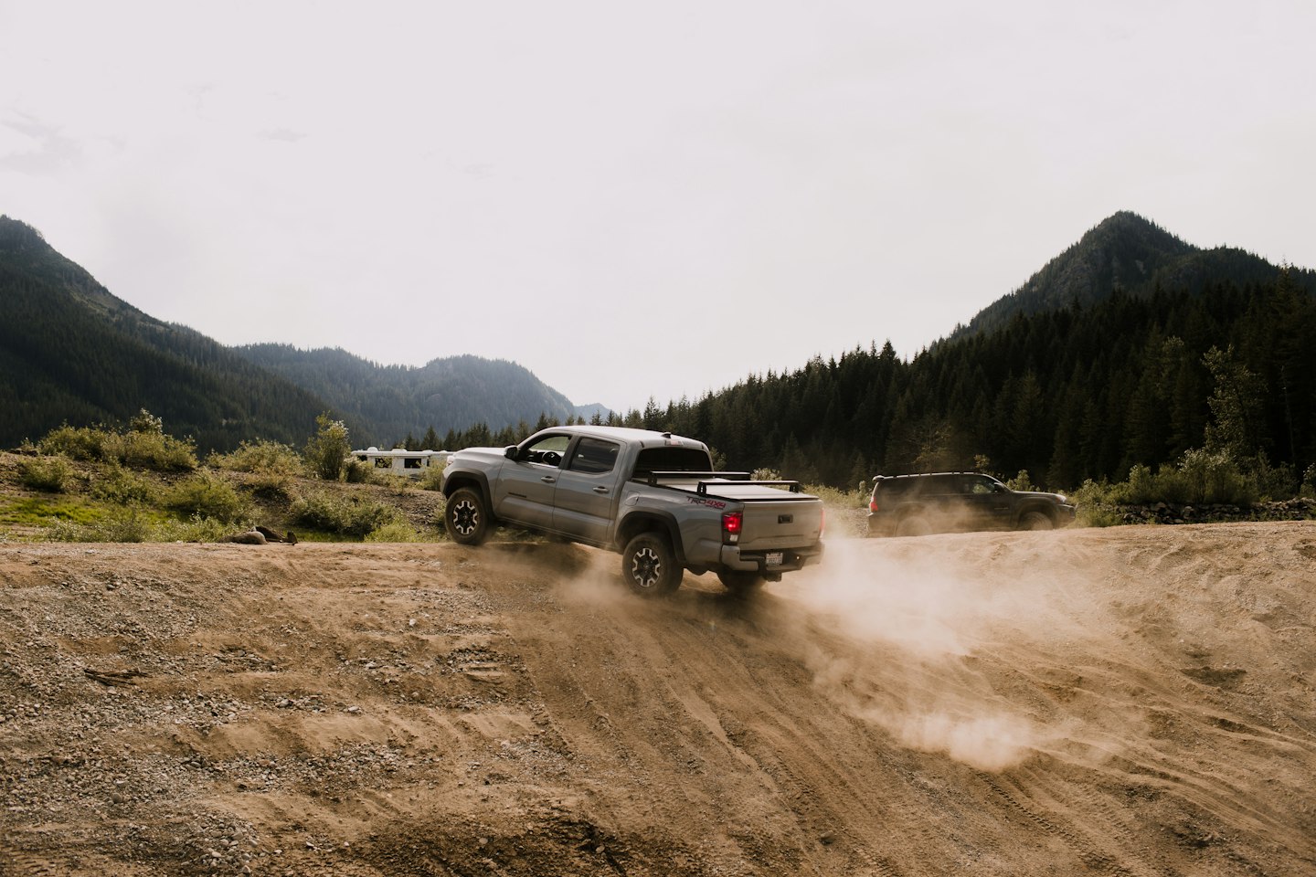 A silver pickup driving along a dusty trail in forested mountains